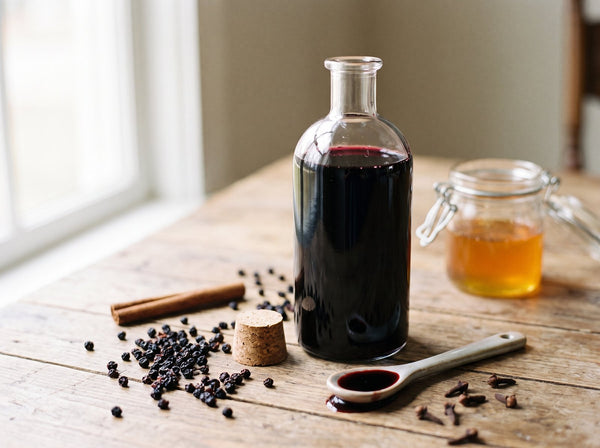A tall clear glass bottle of deep purple-black elderberry syrup on a warm wooden table, cork stopper beside it, a small ceramic spoon holding a glossy pour of the syrup, a scattering of dried elderberries, a cinnamon stick and a f