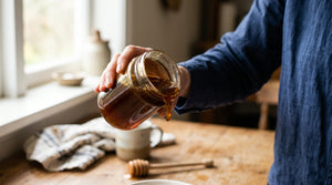 Hand tilting a glass jar of dark Manuka honey toward light, showing thick amber viscosity